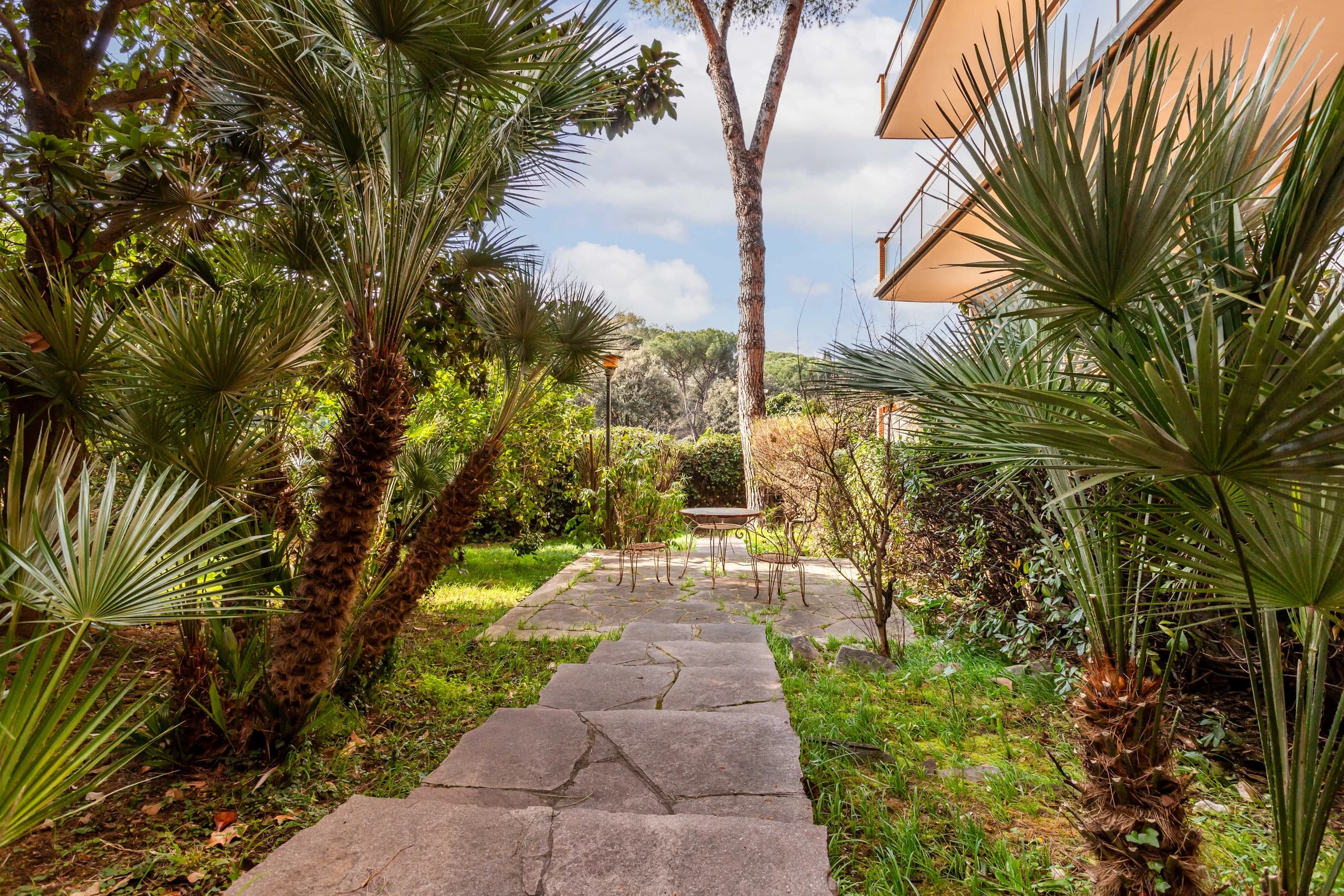 Giardino con palme e camminamento in pietra, con tavolino all'ombra dei pini