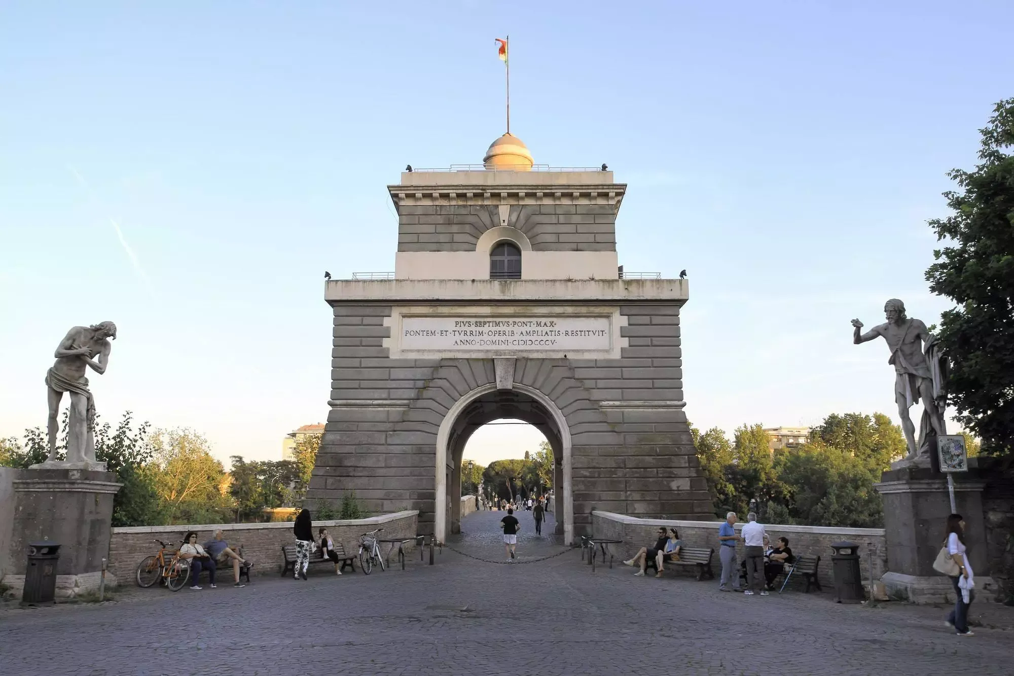 La torretta di Ponte Milvio fra due statue, con persone sedute sulle panchine all'ingresso del ponte